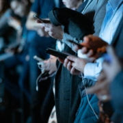Mehrere Personen in formeller Kleidung schauen auf ihre Smartphones, während sie an einer Veranstaltung oder einem Meeting teilnehmen. Die Hände sind deutlich zu sehen, während der Hintergrund unscharf ist.