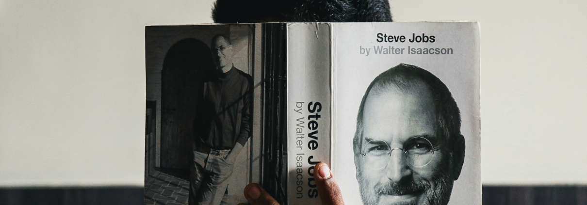 Person hält das Buch Steve Jobs von Walter Isaacson, wobei das Buch das Gesicht von Steve Jobs auf dem Cover zeigt. Der Hintergrund ist unscharf und die Person ist teilweise sichtbar.